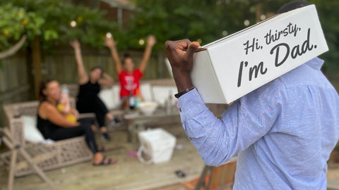Dad Strength Brewing beer box with “Hi, thirsty, I’m Dad” printed on the side, carried over a shoulder at a backyard gathering with friends.